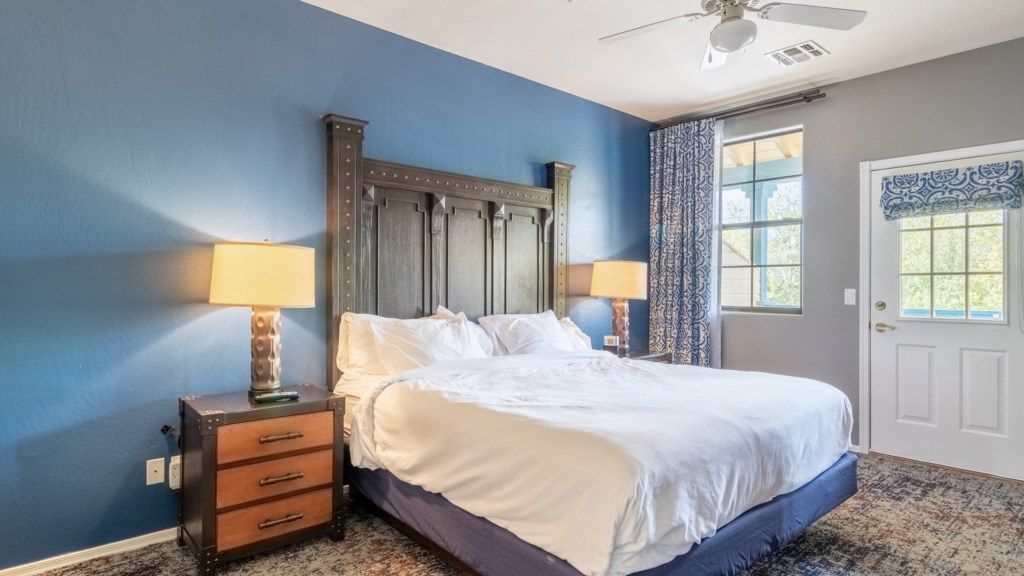 A bedroom with a bed, two bedside lamps, a nightstand, a ceiling fan, and a door with patterned curtains.