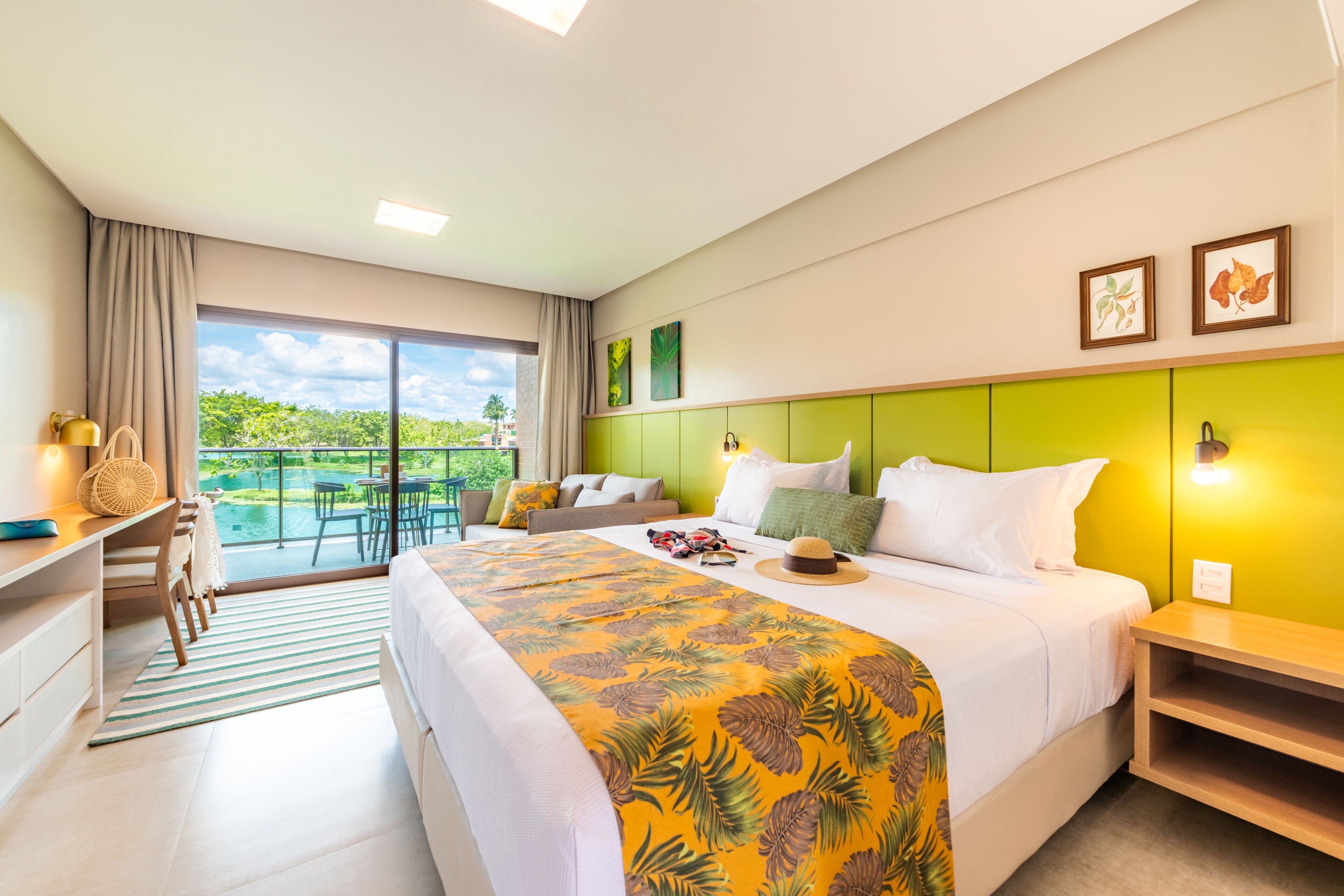 Quarto de hotel com cama, escrivaninha, cadeira, mesa pequena, janela com vista para área verde e varanda.