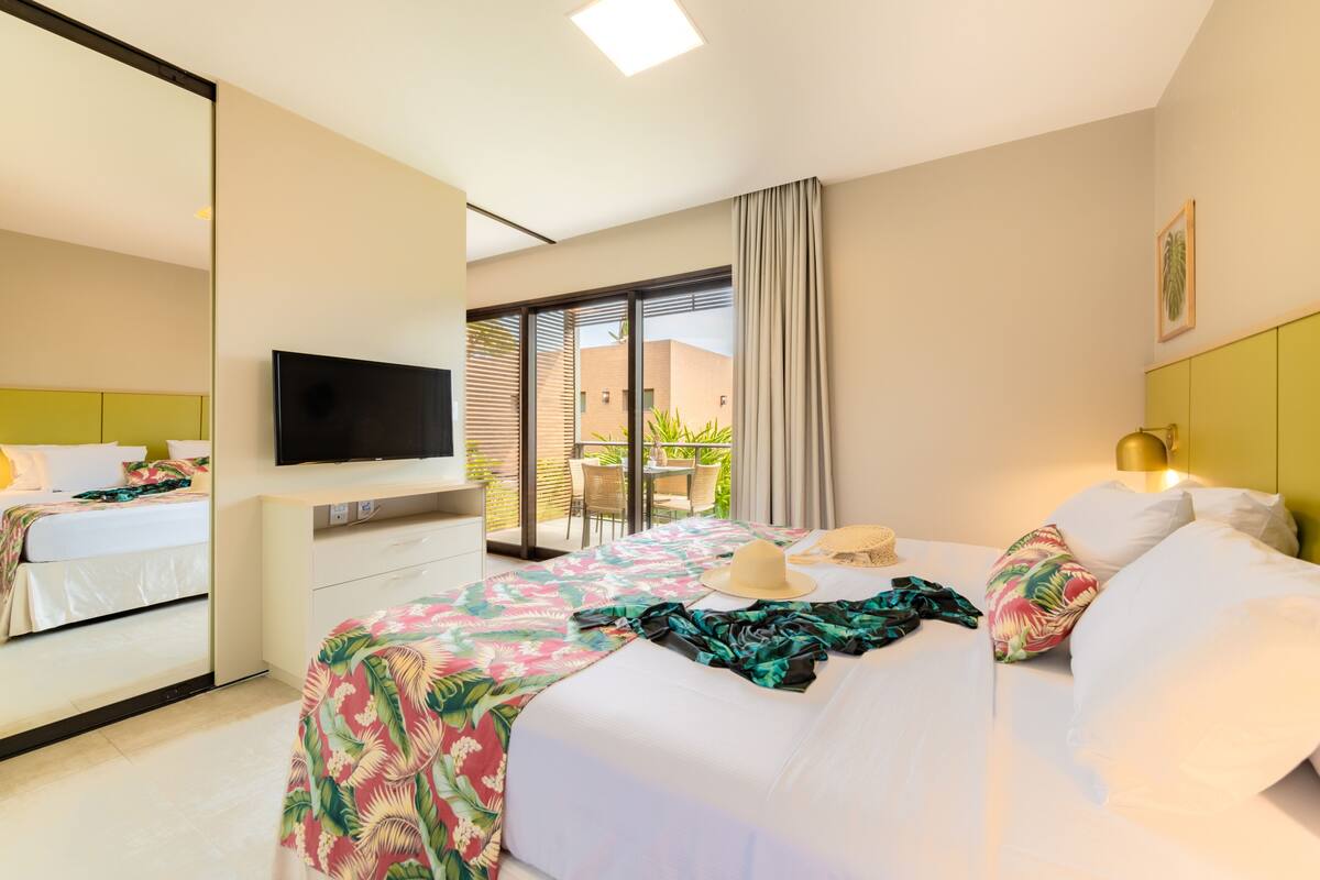 Habitación de hotel con cama, televisor, escritorio y balcón con mesa y sillas.