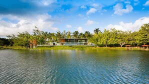 A modern building with a glass facade, situated by a calm lake, surrounded by lush greenery and palm trees. - Reservas dos Lagos - Boa Luz (Laranjeiras)