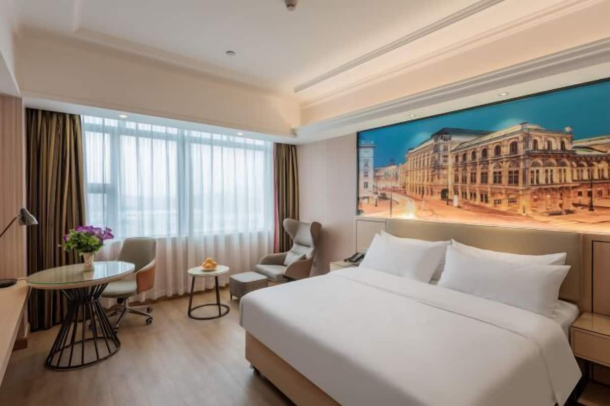 A hotel room with a large bed, a desk, a chair, a small table, and a painting of a building on the wall.