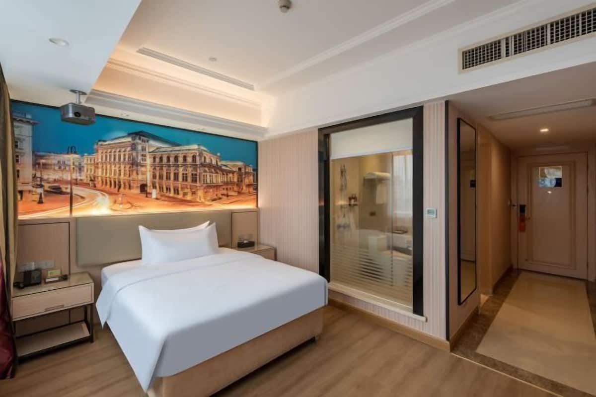 A hotel room with a large bed, a nightstand, a desk, a glass door, and a mural of a cityscape.
