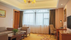 A hotel room with a sofa, armchairs, a desk, a television, and a large window with curtains.