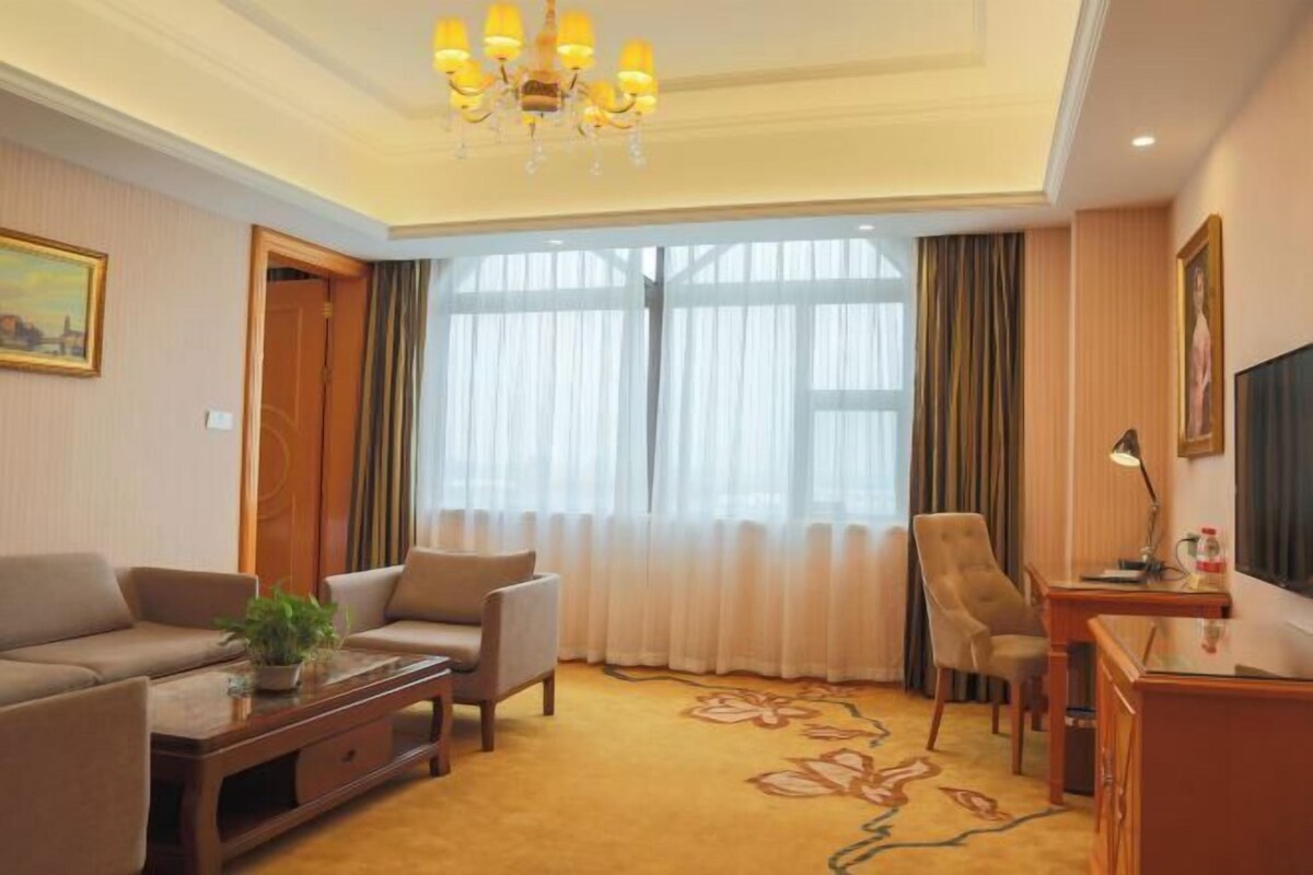 A hotel room with a sofa, armchairs, a desk, a television, and a large window with curtains.