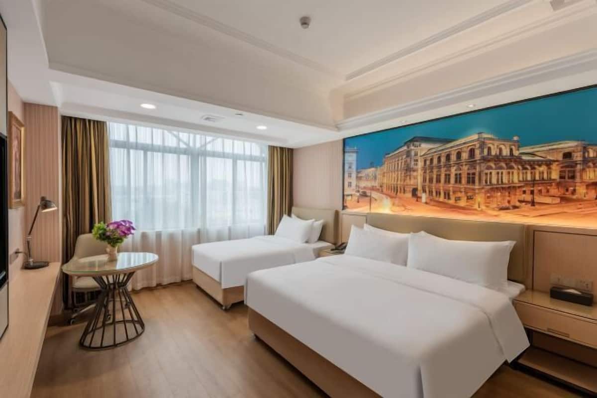 A hotel room with two beds, a small table, a chair, and a painting of a cityscape on the wall.