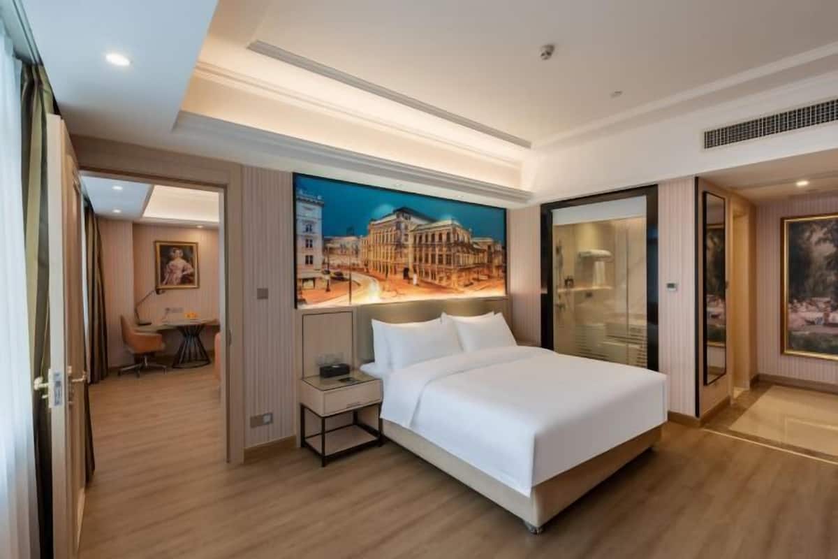 A hotel room with a large bed, a desk with a chair, a painting on the wall, and a view of a building through a glass door.