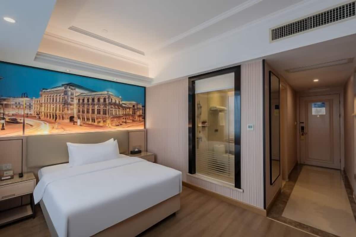 A hotel room with a large bed, a nightstand, a wall-mounted TV, a window with blinds, and a door leading to another room.