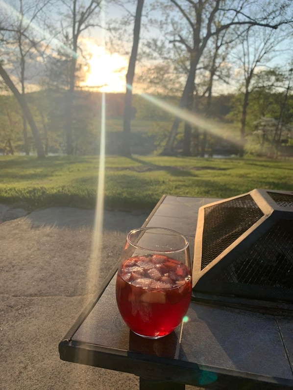 Un verre d’une boisson rouge est posé sur une table à l’extérieur, avec en arrière-plan des arbres et un coucher de soleil.