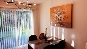 A dining area with a table, chairs, a chandelier, a painting, and a view through a window with blinds.