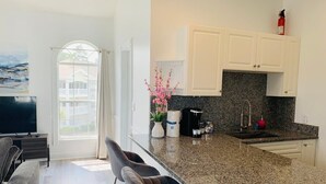 Una cocina moderna con encimeras de granito, gabinetes blancos y horno integrado. Hay un área de comedor con un sofá gris y un televisor de pantalla plana.