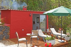 A red building with a stone wall, a glass door, and a patio area with two chairs, a table, and an umbrella. - Casa Antonia en Tijarafe (Tijarafe)