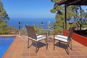 Een terras met rieten stoelen en een tafel, met uitzicht op een zwembad en een prachtig uitzicht over de oceaan.