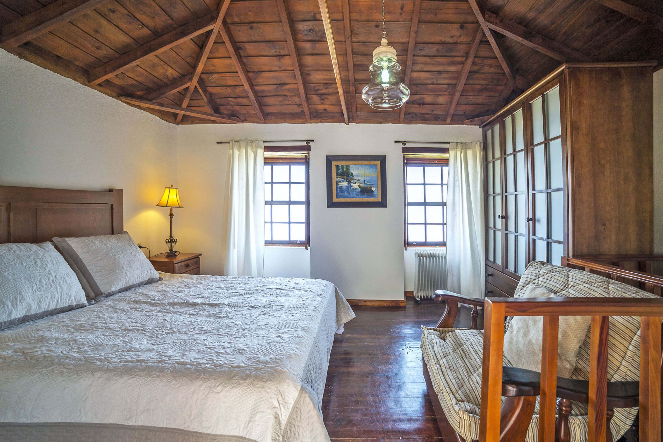 A bedroom with a large bed, a wooden chair, a lamp, and a painting on the wall.