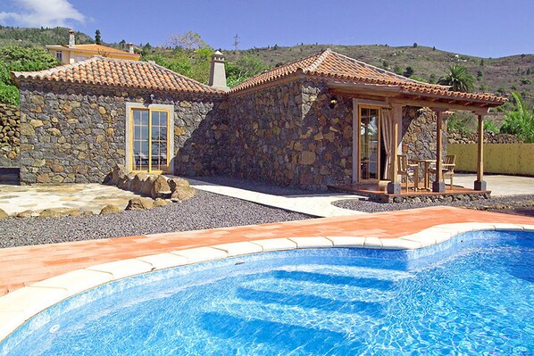 A stone house with a swimming pool, a covered patio with chairs, and a view of hills.