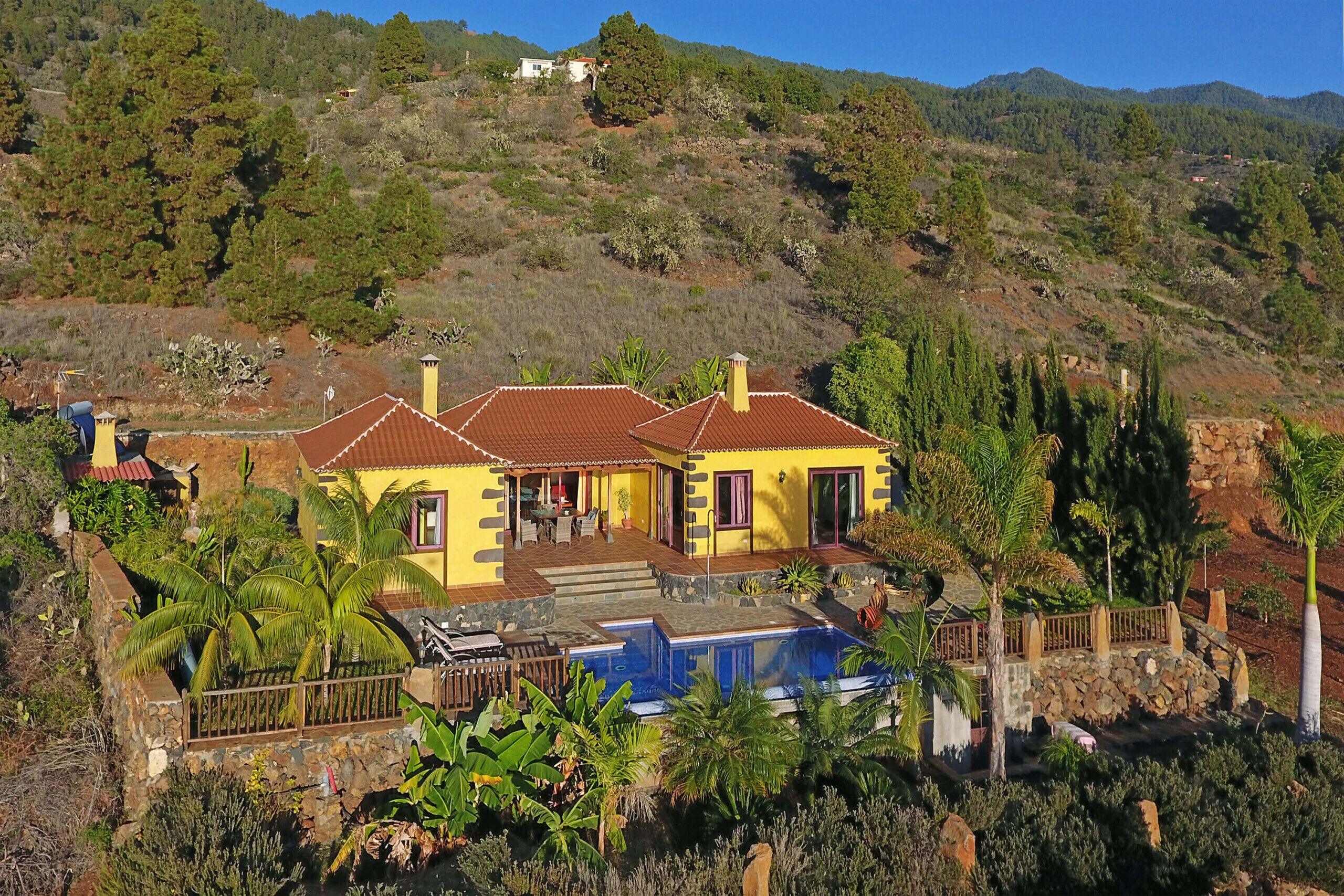 A yellow house with a swimming pool, surrounded by tropical plants and a hilly landscape.
