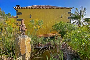 A yellow building with a tiled roof, surrounded by lush greenery and a small pond.