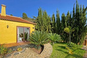 A yellow building with a tiled roof, a stone pathway, and a variety of plants including a palm and a fern.