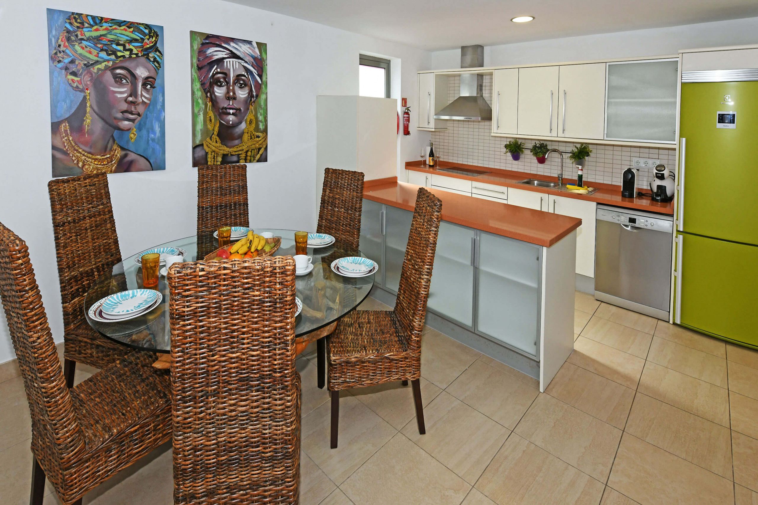 Un coin repas avec des chaises en osier et une table en verre, une cuisine avec des meubles blancs et un réfrigérateur vert, et deux tableaux accrochés au mur.