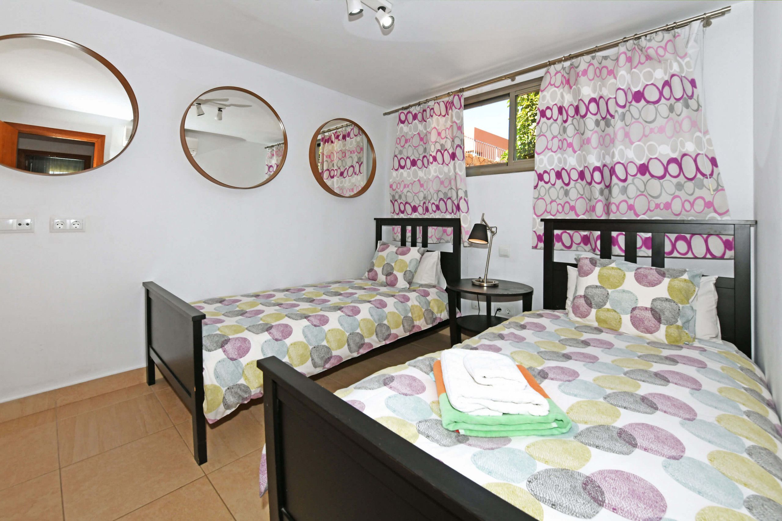 A bedroom with two single beds, a small table, a lamp, and a window with curtains.