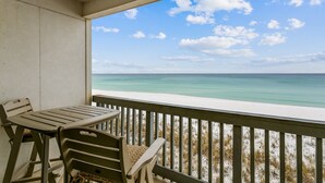 A balcony with a table and chairs, overlooking a beach with clear turquoise water and a sandy shore. - Shore Beats Work! Gulf Front! Community Pool! (Penscola Beach)