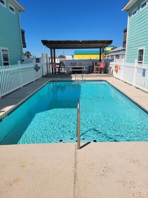 Une piscine aux eaux d’un bleu limpide, entourée d’une clôture blanche, avec un poste de surveillance pour les sauveteurs et un espace de repos abrité.