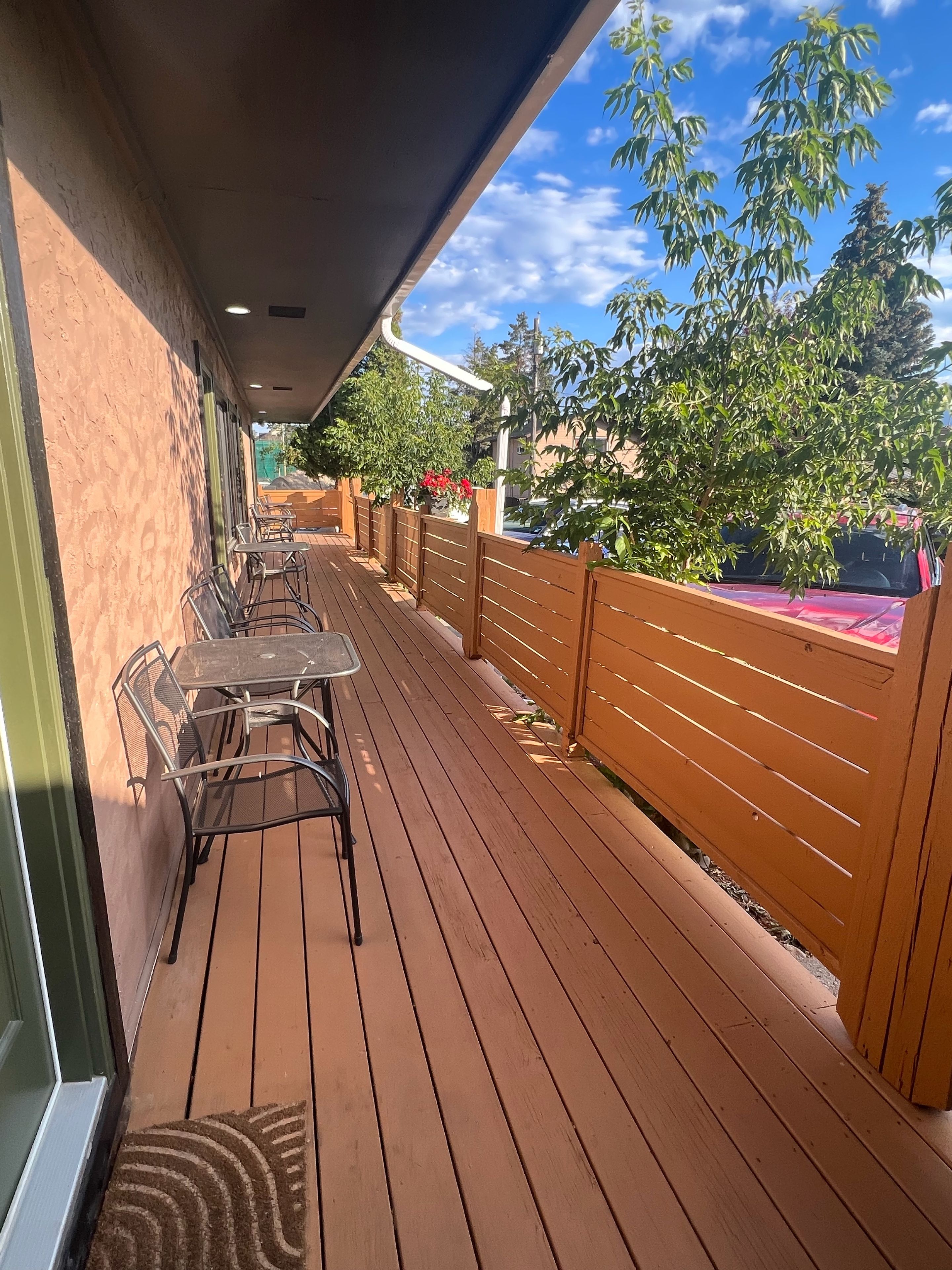 An outdoor patio with wooden flooring, metal tables and chairs, and a view of trees and a car in the background.