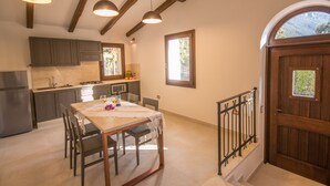 A kitchen with wooden beams, a dining table with chairs, and a view of the outdoors through a window. - Villa Capelvenere (Corbezzolo) (Dorgali)