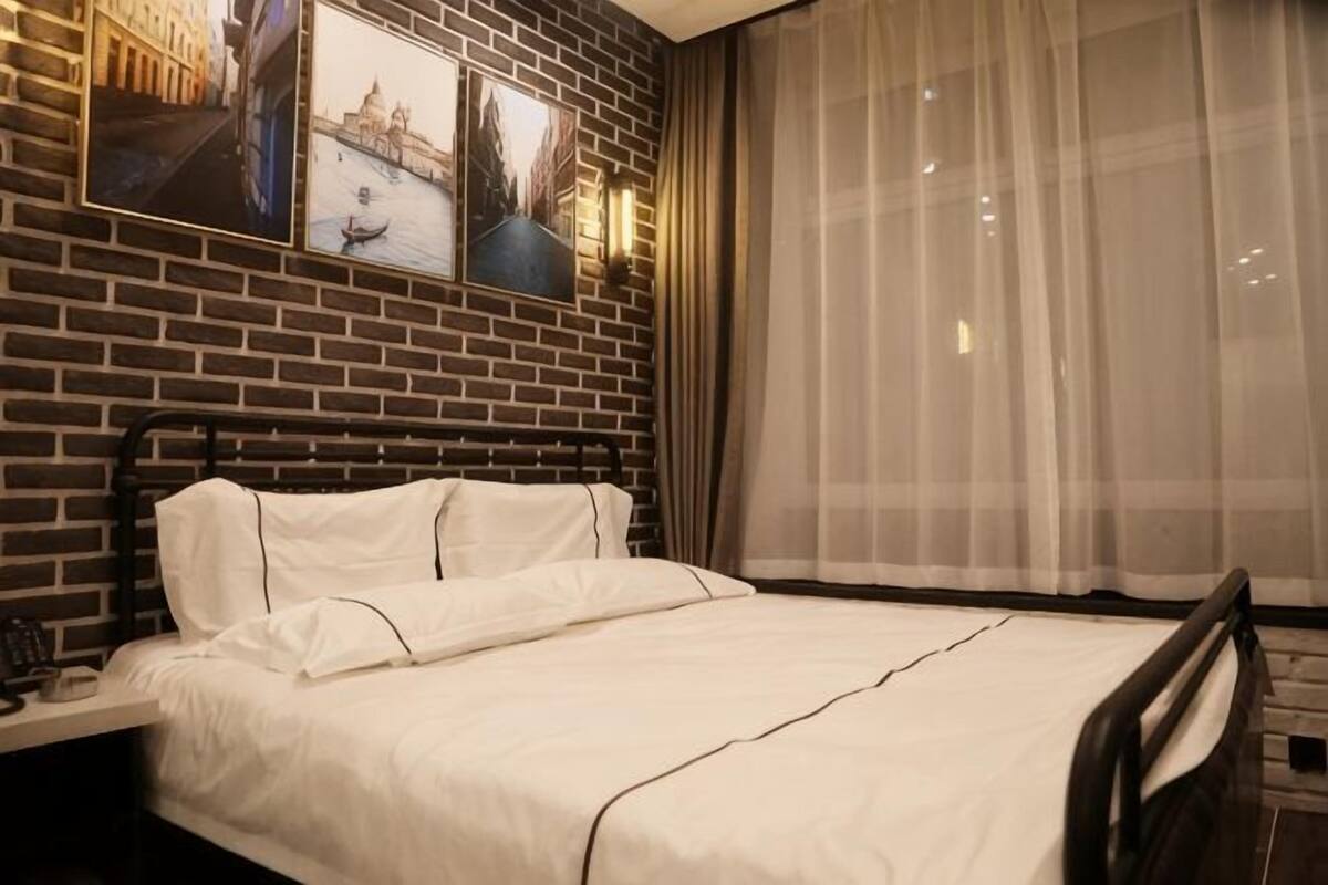 A hotel room with a brick wall, a bed with white linens, a nightstand, and a window with sheer curtains.