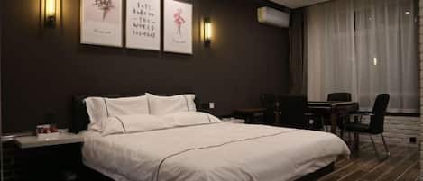 A modern hotel room with a large bed, bedside tables, a desk, and a chair. There are wall-mounted lights and a large window with curtains.