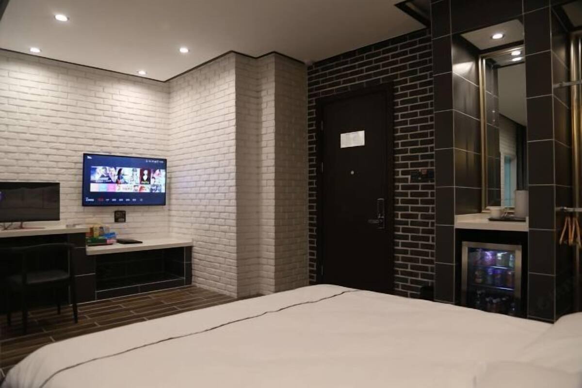 A modern hotel room with a brick accent wall, a flat-screen TV, a desk, and a minibar.