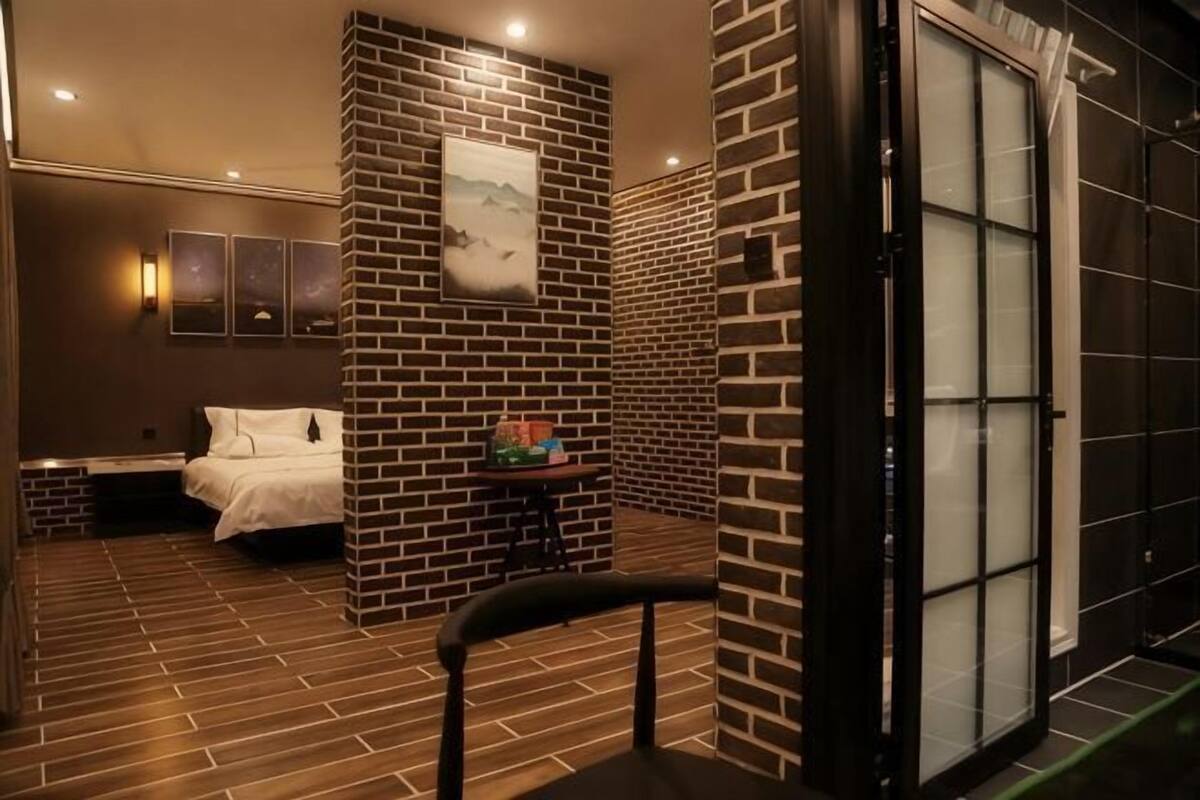 A hotel room with a brick accent wall, a bed, a small table with a chair, and a large window.