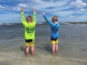 Two children standing in shallow water with their arms raised, wearing long-sleeve shirts and shorts.