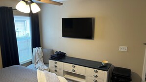A hotel room with a bed, a desk with drawers, a television on the wall, a ceiling fan, and a window with curtains.