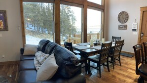 A cozy living room with a leather sofa, wooden dining table, and chairs. Large windows offer a view of a snowy deck and trees. - Fully Stocked Family Friendly Cabin Near Breckenridge (Fairplay)