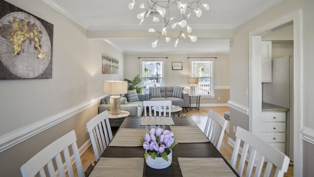 A dining area with a wooden table, white chairs, and a vase of purple flowers. There is a gray sofa and a white coffee table in the background.
