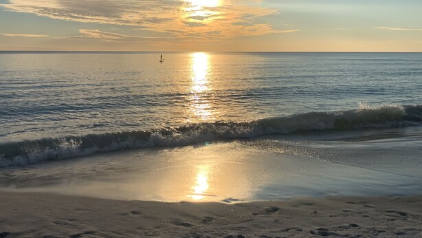 Una playa con mar tranquilo, una persona remando y un atardecer.