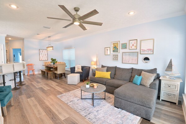 A living room with a gray sofa, a coffee table, and a ceiling fan. There are framed pictures on the wall and a lamp on a side table.