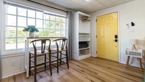 Une pièce avec un plancher en bois, une fenêtre avec des stores, un bureau avec une chaise, une porte jaune et une petite table blanche avec une lampe.