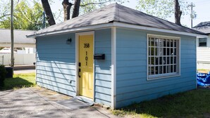 Un petit bâtiment d’un seul étage, avec une porte jaune et une fenêtre grillagée, situé dans une zone herbeuse, avec des arbres en arrière-plan.