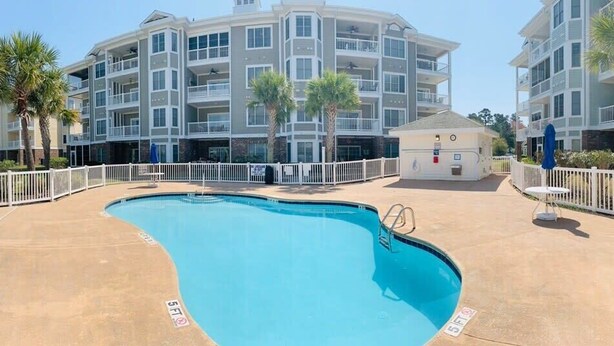 A swimming pool with a white fence, palm trees, and a multi-story building in the background.