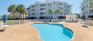 A swimming pool with a white fence, palm trees, and a multi-story building in the background.
