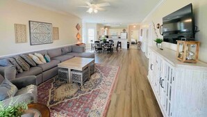A spacious living room with a gray sofa, a wooden coffee table, and a patterned rug. A dining area with a white table and chairs is visible in the background.