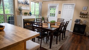 A dining area with a wooden table and chairs, a fireplace, and a view of greenery through large windows. - MTN View Cottage Getaway Stunning Fall Colors Private Bathroom per bedroom (Sevierville)