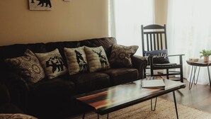 A living room with a dark brown leather sofa, a wooden coffee table, and a woven rug. There are wall-mounted animal-themed artwork and a ceiling fan. - MTN View Cottage Getaway Stunning Fall Colors Private Bathroom per bedroom (Sevierville)