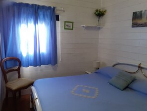 Una habitación con una cama, una silla, un ventanal con cortinas azules, un pequeño estante con un florero y un cuadro en la pared.