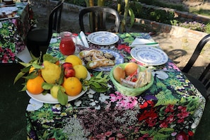 Une table dressée à l’extérieur, recouverte d’une nappe à motifs floraux, avec des assiettes de fruits, du pain et un panier de petits pains.