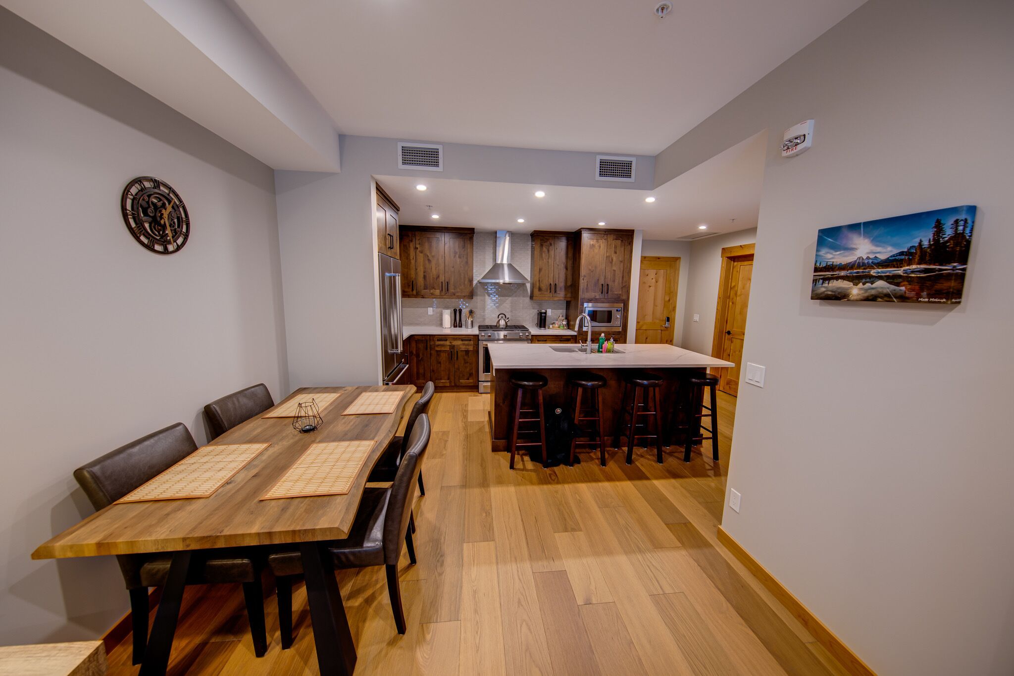 A modern kitchen with wooden cabinets, a central island with stools, and a dining table with chairs.