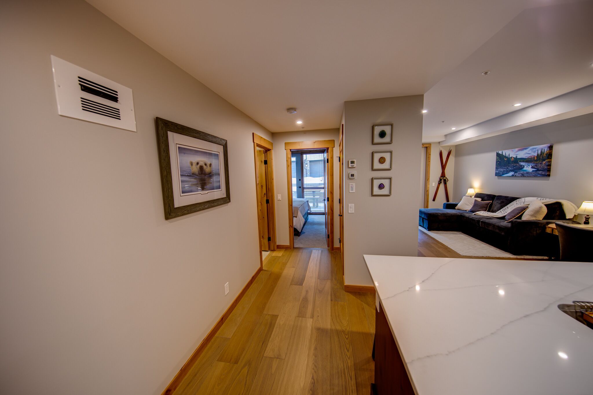 A hallway with wooden flooring, a framed picture on the left, and a dark-colored sofa with a lamp on the right.