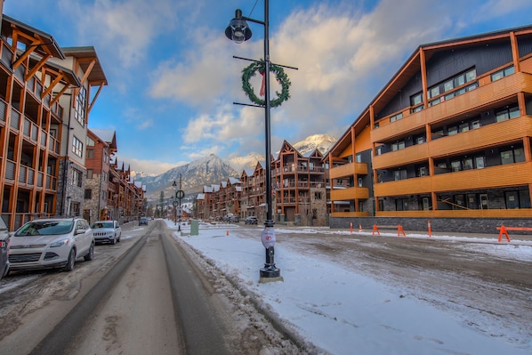 A snow-covered street with multi-story buildings, including a hotel with balconies and a mountain backdrop.