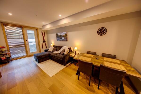 Ein modernes Wohnzimmer mit einer Couch, einem Esstisch mit Stühlen, einem Kamin und einem großen Fenster mit Jalousien.